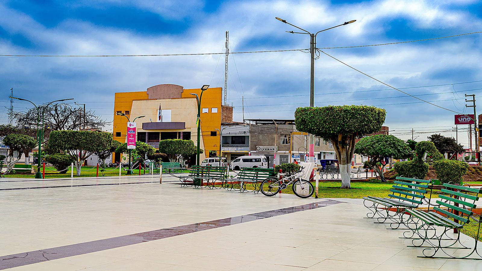Plaza principal de Pueblo Nuevo iluminada por la noche, con pileta ornamental y áreas verdes.